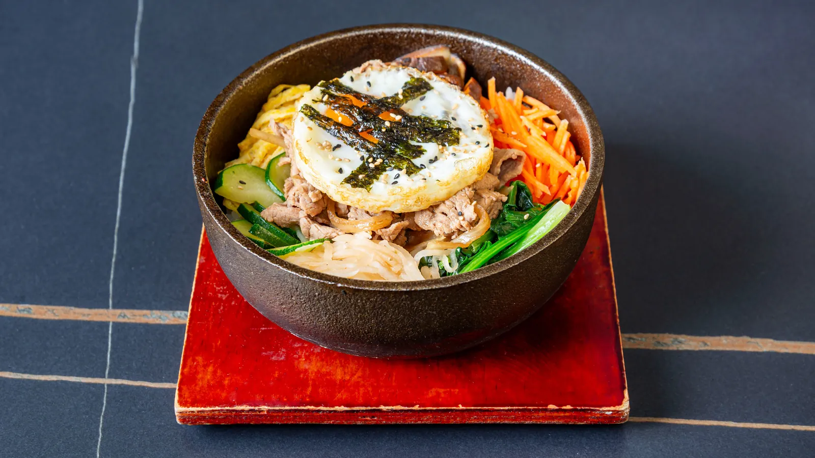 Bibimbap au bœuf et légumes dans un bol en fonte, sur un dessous de plat rouge