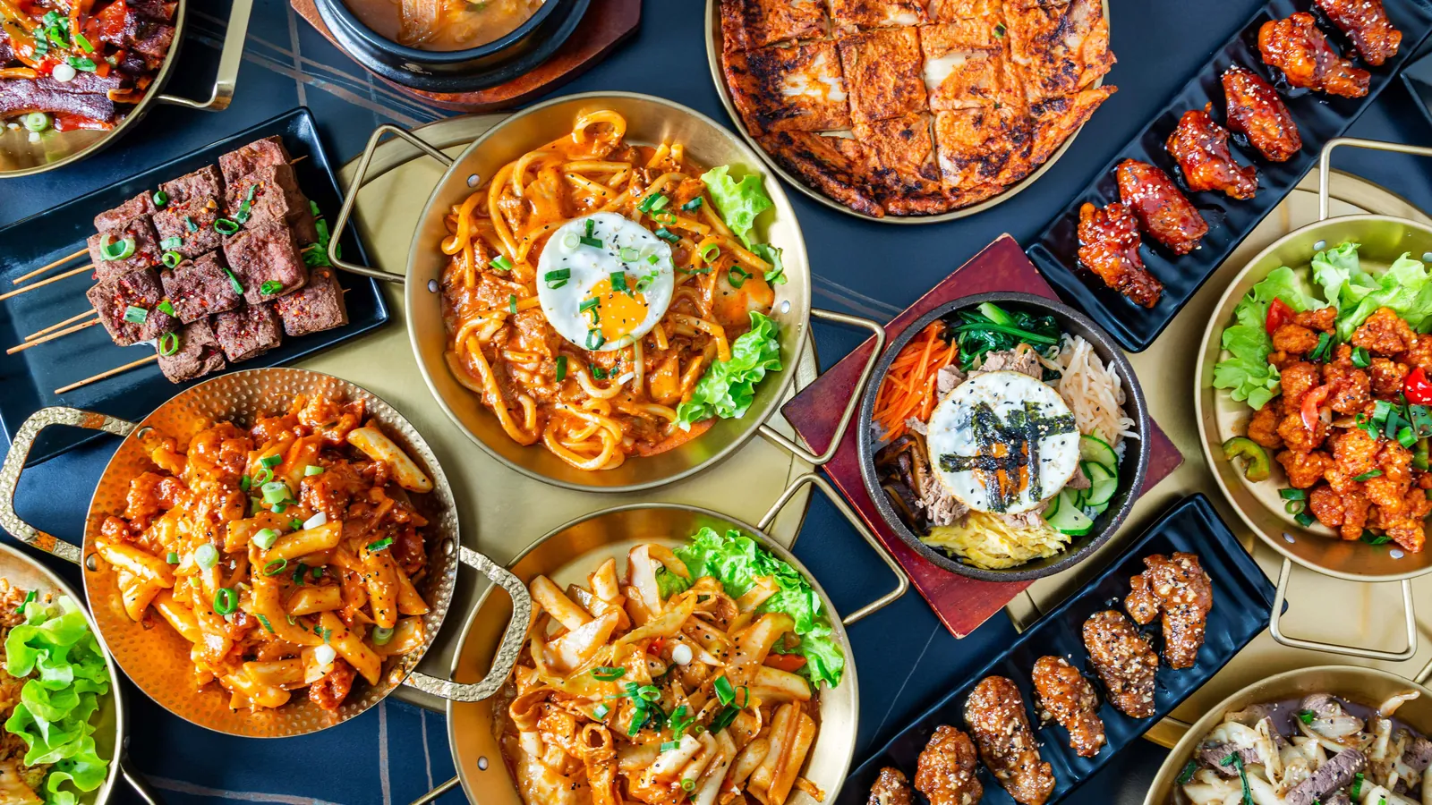 Vue du dessus d'une table de plats coréens : brochettes, bibimbap, galette, ailes de poulet et pâtes de riz