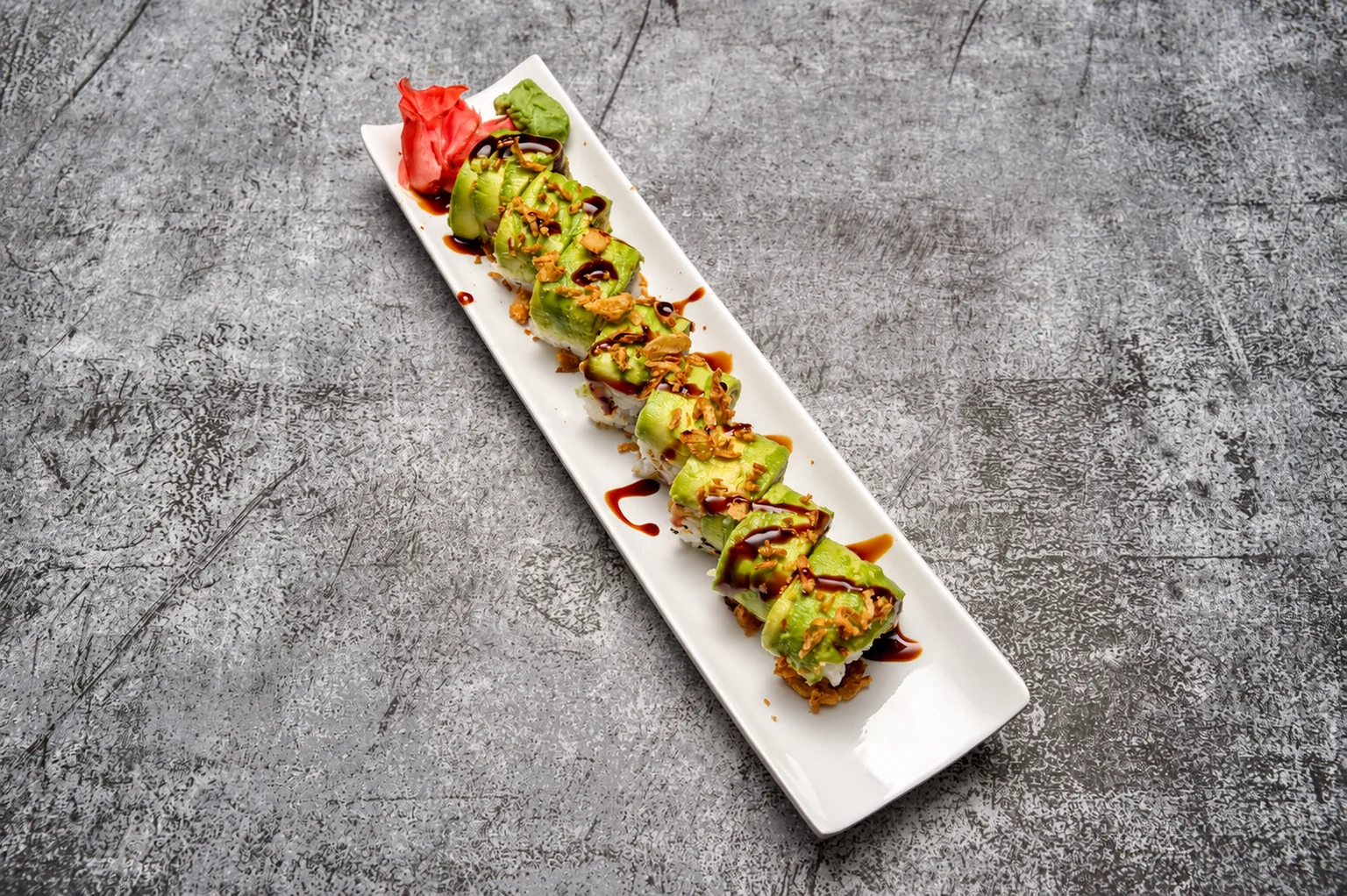 Rolls recouverts de tranches d'avocat et de sauce sur une longue assiette blanche, fond de béton gris.